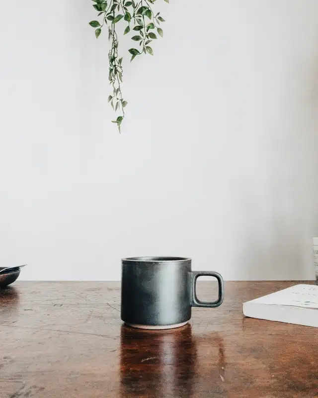 Caneca de cerâmica escura com alça quadrada sobre uma mesa de madeira, com uma planta pendurada e uma parede branca ao fundo.