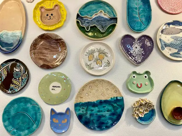 Parede branca coberta de pratinhos de cerâmica esmaltados, de formatos e cores variados, como um gato, um sapo e um coração roxo.
