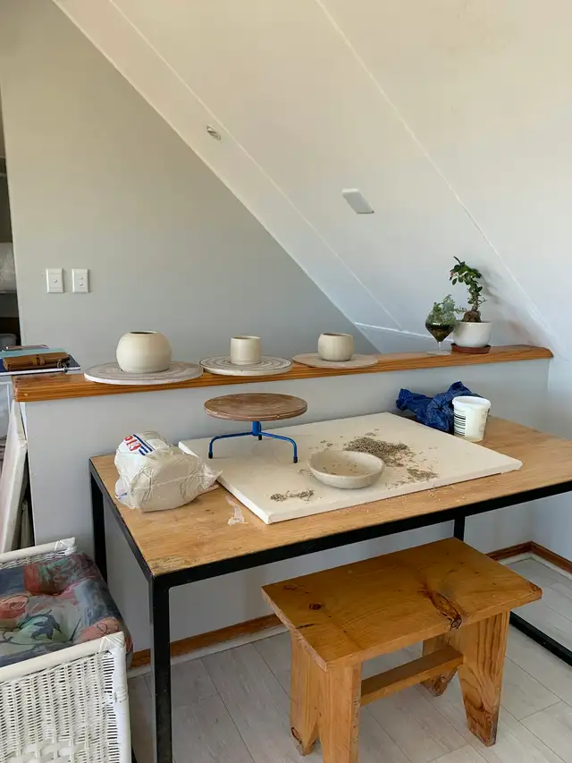 Cantinho de ateliê de cerâmica: bancada com um bloco de argila, um torno de mesa e uma tigela em modelagem; na prateleira ao fundo, três peças de argila crua e uma plantinha.