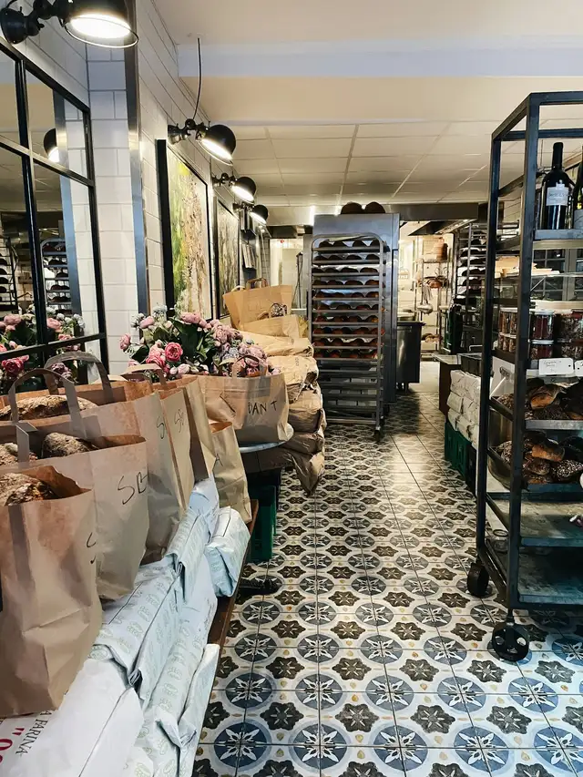 Corredor de uma padaria com sacolas de papel cheias de pães à esquerda, um carrinho de assadeiras com pãezinhos ao fundo e o piso de ladrilhos estampados.
