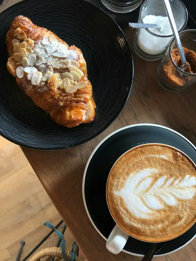 Vista de cima de um croissant com amêndoas laminadas num prato preto e de um cappuccino com arte no leite, ao lado de potes de açúcar, sobre uma mesa de madeira.